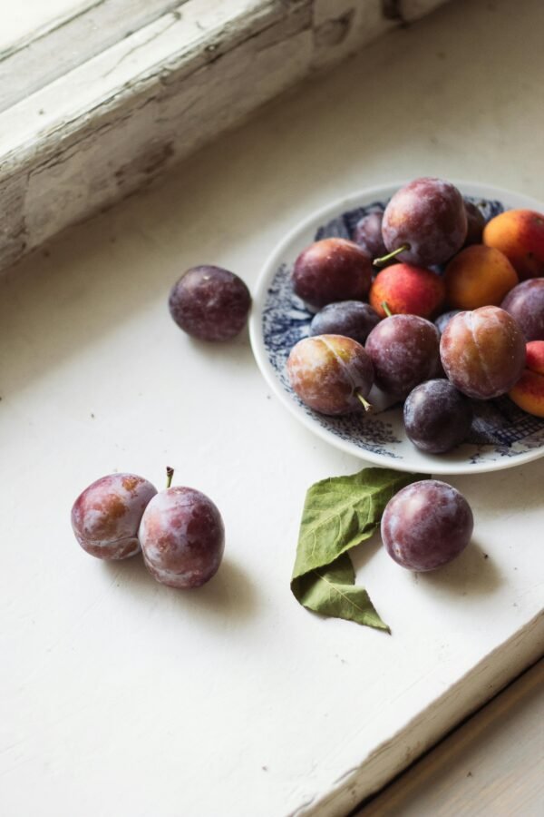 A rustic display of fresh plums on a plate by a sunlit windowsill, creating a cozy and inviting scene.