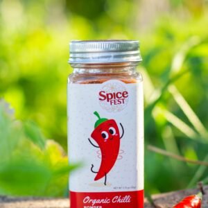 Close-up of organic chilli powder in a labeled jar with fresh peppers, outdoors.