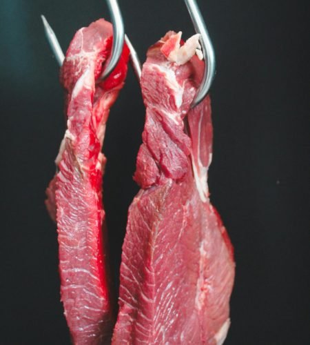 Two raw beef cuts hanging on metal hooks against a dark background.